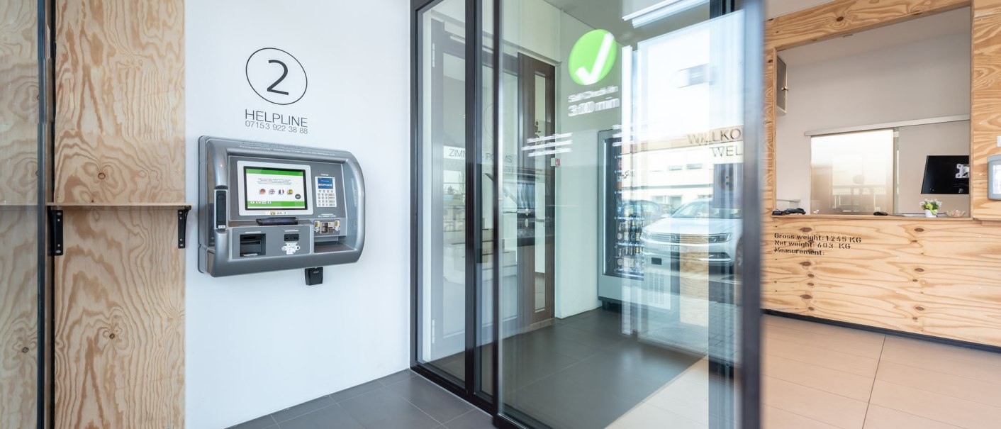 Modern entrance area with self-service terminal on the wall, glass door and wooden reception desk., © a2 HOTELS Modern entrance area with self-service terminal on the wall, glass door and wooden reception desk., © a2 HOTELS