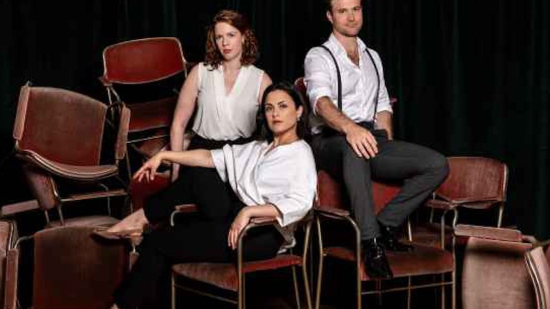 Drei Personen in eleganter Kleidung posieren auf gestapelten, roten Stühlen vor einem dunklen Vorhang., © Internationale Hugo-Wolf-Akademie Drei Personen in eleganter Kleidung posieren auf gestapelten, roten Stühlen vor einem dunklen Vorhang., © Internationale Hugo-Wolf-Akademie