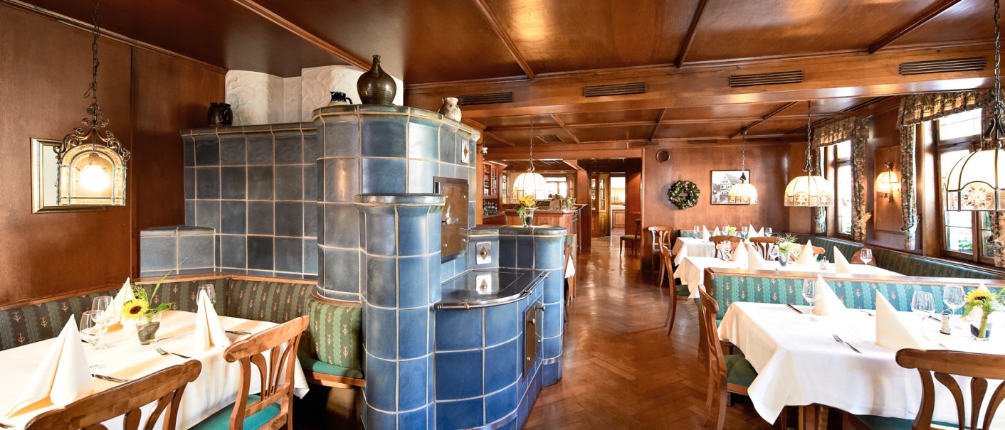 Gemütliches Restaurant mit Holzmöbeln, blauen Kachelofen und gedeckten Tischen. Traditionelle Atmosphäre mit warmem Licht und Dekorationen., © Hirsch Hotel