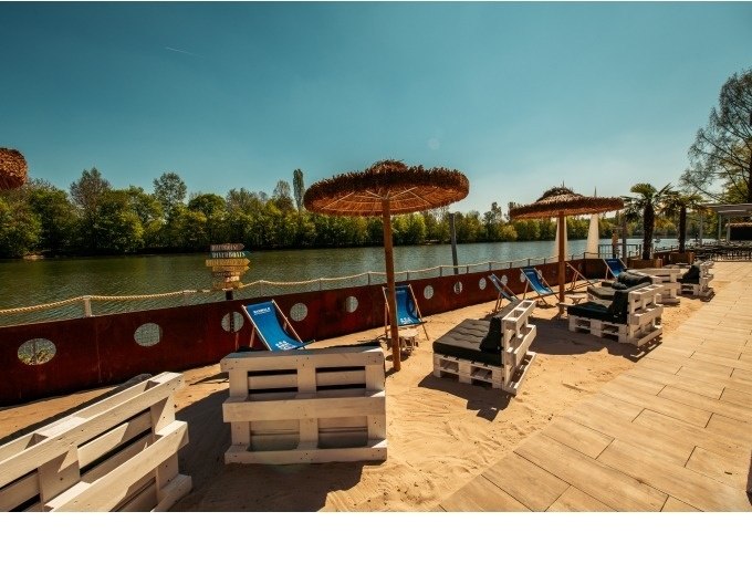 A sandy beach area with blue sun loungers and thatched umbrellas on the banks of a river, surrounded by trees., © Cool-Tours StattReisen A sandy beach area with blue sun loungers and thatched umbrellas on the banks of a river, surrounded by trees., © Cool-Tours StattReisen