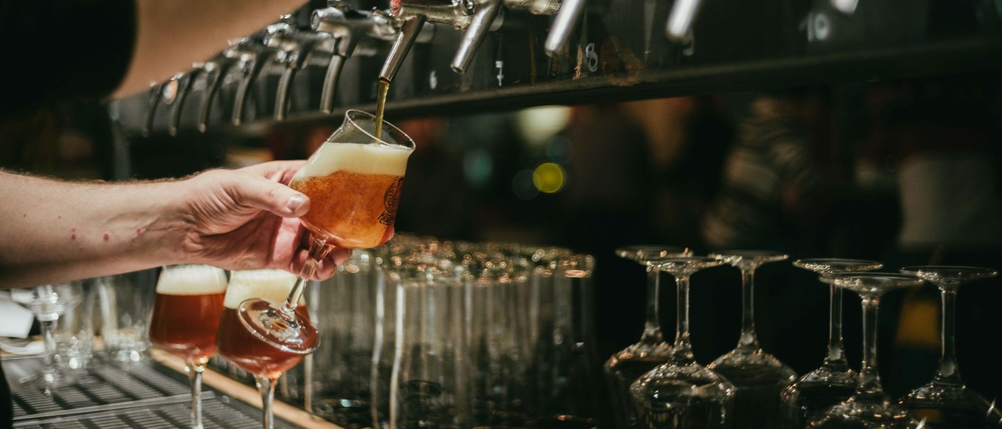 A bartender draws beer from a tap into a glass. More glasses are ready. The scene takes place at a bar., © Kraftpaule GmbH A bartender draws beer from a tap into a glass. More glasses are ready. The scene takes place at a bar., © Kraftpaule GmbH