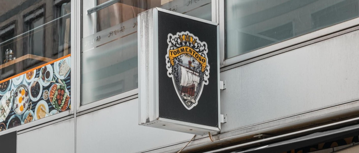 Ein Schild mit dem Logo von Kap Tormentoso hängt an einem modernen Gebäude. Es zeigt ein Segelschiff und dekorative Elemente., © Stuttgart Marketing GmbH, Sarah Schmid