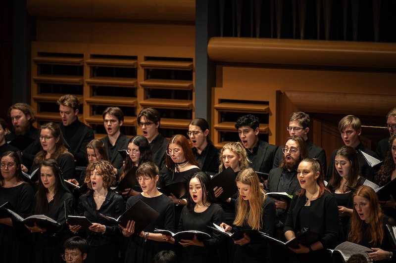 Ein Chor in schwarzer Kleidung singt auf einer B&uuml;hne. Die S&auml;ngerinnen und S&auml;nger halten Notenbl&auml;tter in den H&auml;nden., &copy; Staatliche Hochschule f&uuml;r Musik und Darstellende Kunst