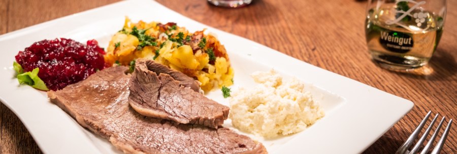 Boiled beef with potato gratin, cranberries and creamed horseradish on a white plate. A glass of wine in the background., © Weingut Zaiß