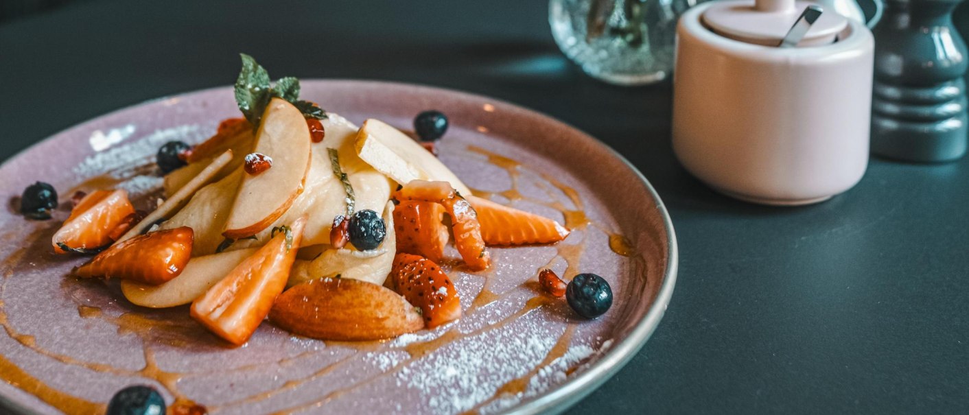 Ein Teller mit geschnittenen Früchten, Beeren und Sirup. Daneben eine Zuckerdose und ein Salzstreuer auf einem dunklen Tisch., © Lupin, Stuttgart Ein Teller mit geschnittenen Früchten, Beeren und Sirup. Daneben eine Zuckerdose und ein Salzstreuer auf einem dunklen Tisch., © Lupin, Stuttgart