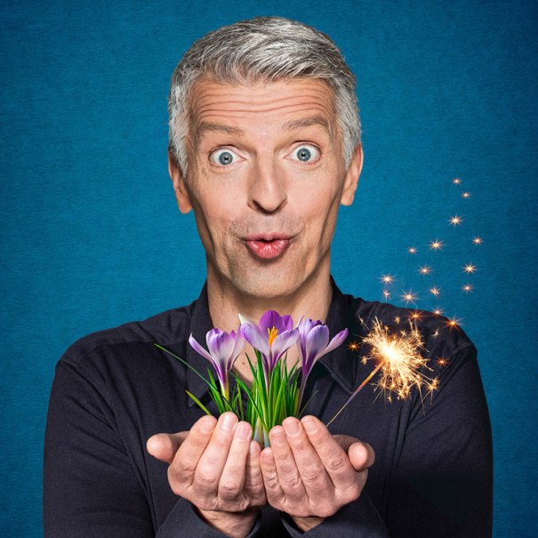 Ein Mann mit grauen Haaren hält Krokusse und eine Wunderkerze in den Händen vor blauem Hintergrund., © Renitenztheater Stuttgart e.V. Ein Mann mit grauen Haaren hält Krokusse und eine Wunderkerze in den Händen vor blauem Hintergrund., © Renitenztheater Stuttgart e.V.