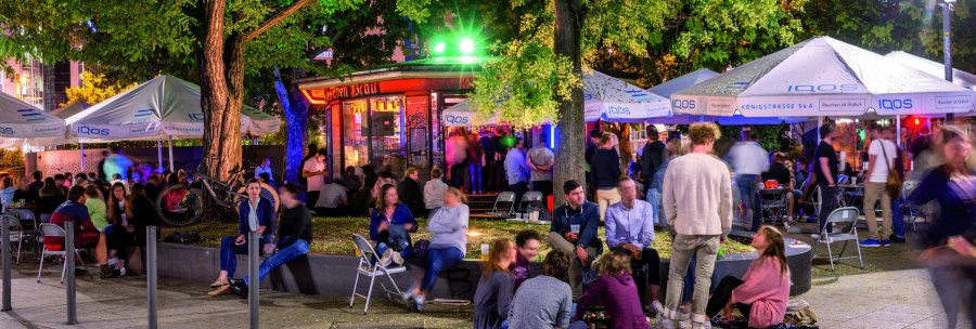 Lively outdoor area with people under illuminated umbrellas and trees. Some are sitting on chairs, others on the ground. Colorful lights in the background., © TMBW, Gregor Lengler
