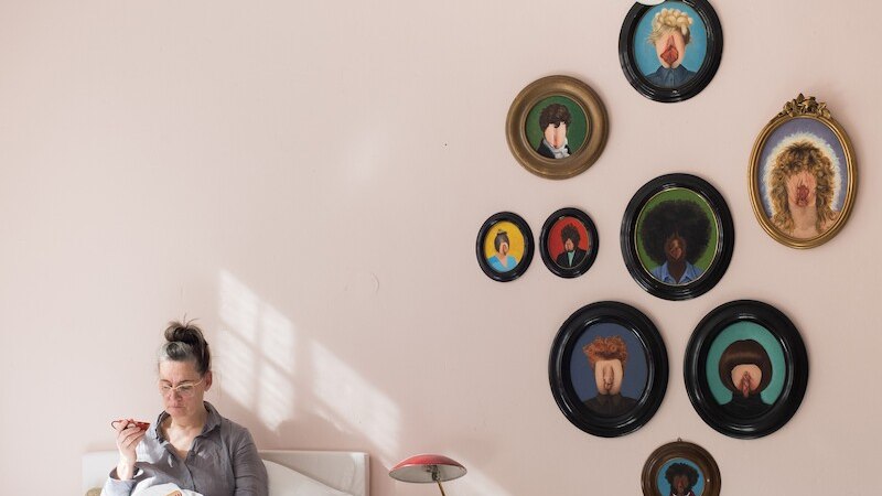 Person sitzt im Bett und liest ein Buch. An der rosa Wand hängen ovale Porträts. Eine Lampe steht auf einem Karton neben dem Bett., © Theater Rampe