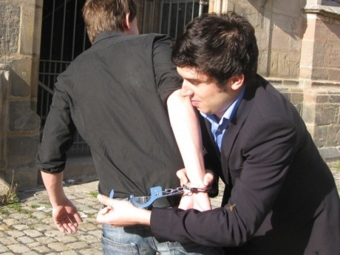 A man handcuffs another man outside. Both are wearing dark clothing. A building can be seen in the background., © Cool-Tours StattReisen A man handcuffs another man outside. Both are wearing dark clothing. A building can be seen in the background., © Cool-Tours StattReisen