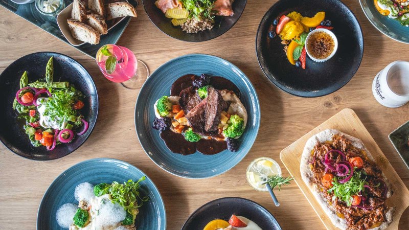 Ein Tisch mit verschiedenen Gerichten: Fleisch mit Gemüse, Salate, Burger mit Pommes, Brot und Desserts. Getränke sind ebenfalls zu sehen., © SatzDrei