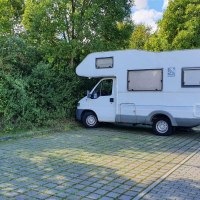 Ein Wohnmobil steht auf einem gepflasterten Stellplatz neben einer Ladestation, umgeben von Bäumen und einem Gebäude im Hintergrund., © Stadt Schorndorf Ein Wohnmobil steht auf einem gepflasterten Stellplatz neben einer Ladestation, umgeben von Bäumen und einem Gebäude im Hintergrund., © Stadt Schorndorf