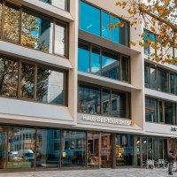 Moderne Glasfassade des Haus des Tourismus mit Spiegelungen von Bäumen und umliegenden Gebäuden. Menschen stehen vor dem Eingang., © SMG, Sarah Schmid Moderne Glasfassade des Haus des Tourismus mit Spiegelungen von Bäumen und umliegenden Gebäuden. Menschen stehen vor dem Eingang., © SMG, Sarah Schmid