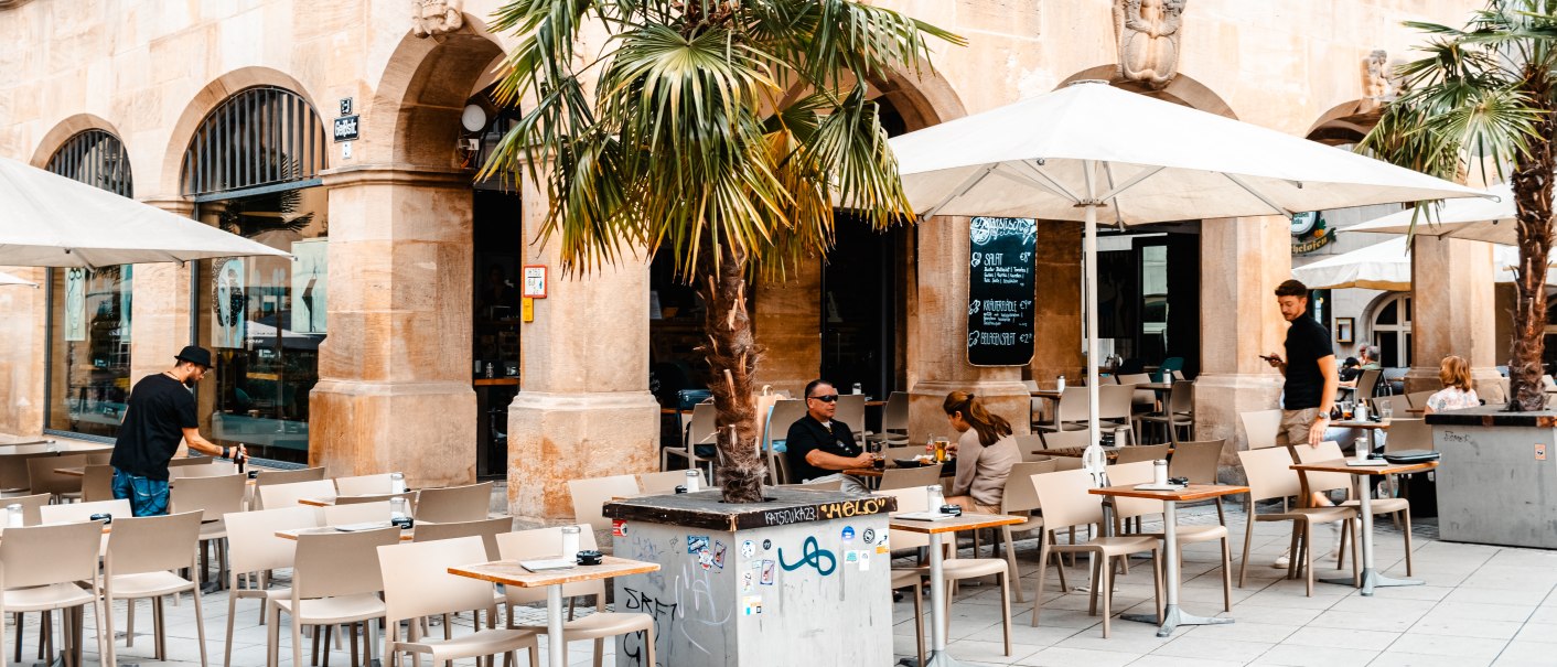 Außenbereich eines Cafés mit Tischen, Stühlen und Sonnenschirmen vor einem historischen Gebäude. Menschen sitzen und bedienen sich., © SMG Stuttgart Marketing GmbH - Sarah Schmid
