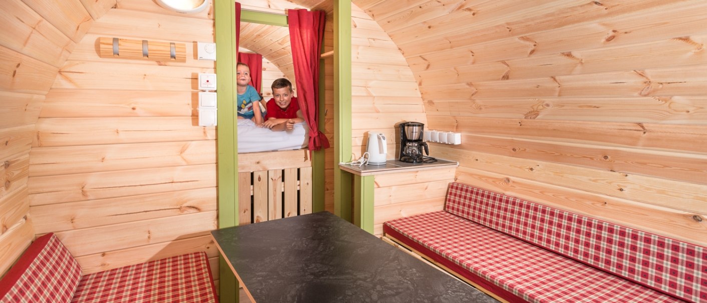 Innenansicht eines gemütlichen Holzfasses mit Tisch, gepolsterten Bänken und zwei Kindern auf einem Hochbett. Kaffeemaschine und Wasserkocher vorhanden., © Remsfässer