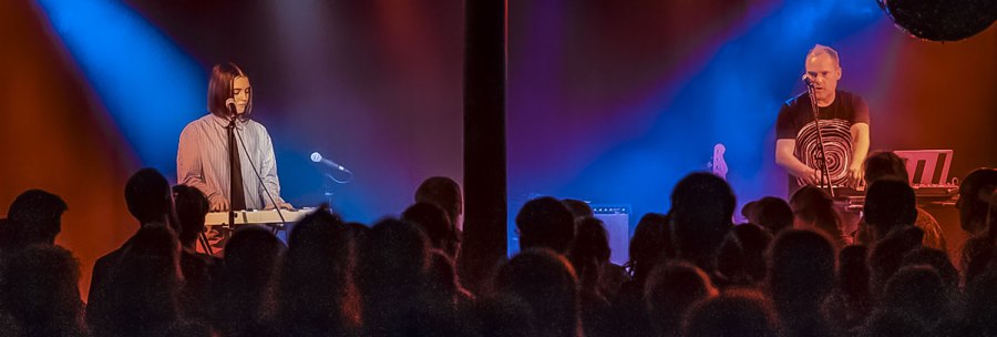 Two musicians on a stage play keyboards. Colorful lights in blue and red illuminate the scene. An audience looks on., © Martin Schniz Two musicians on a stage play keyboards. Colorful lights in blue and red illuminate the scene. An audience looks on., © Martin Schniz