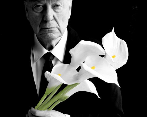 An older man in a suit and cap holds a bouquet of white calla lilies against a black background. The expression is serious and thoughtful., &copy; Theaterhaus Stuttgart e.V.