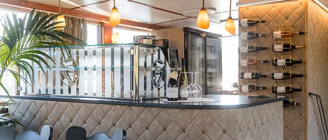 Elegante Bar mit gepolstertem Tresen, Weinflaschen an der Wand und stilvoller Beleuchtung. Eine Palme und Gl&auml;ser erg&auml;nzen das Ambiente., &copy; NeckarFlair MS Wilhelma Betriebsgesellschaft mbH