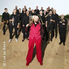 Eine Blasmusikgruppe steht im Freien, ein Mann im roten Anzug mit Trompete im Vordergrund. Die Gruppe trägt schwarze Kleidung., © links im Bild Eine Blasmusikgruppe steht im Freien, ein Mann im roten Anzug mit Trompete im Vordergrund. Die Gruppe trägt schwarze Kleidung., © links im Bild