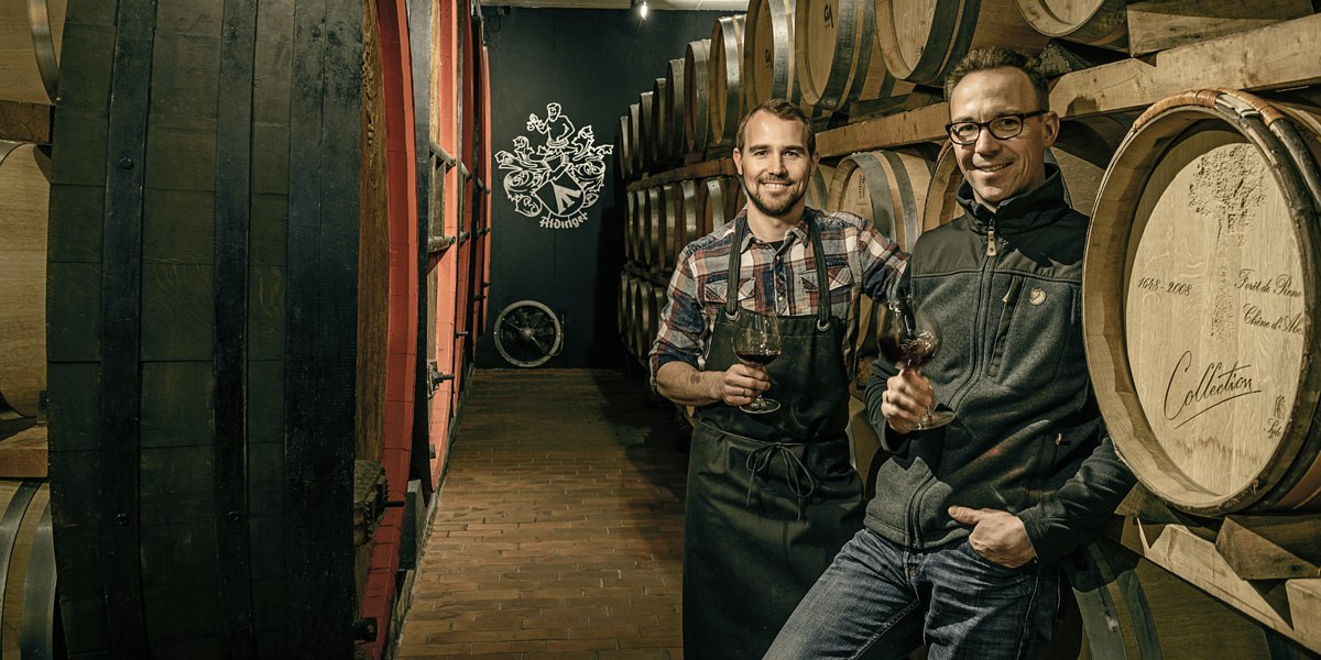 Two men in a wine cellar with wooden barrels, holding wine glasses. In the background is a logo on the wall., © Weingut Aldinger