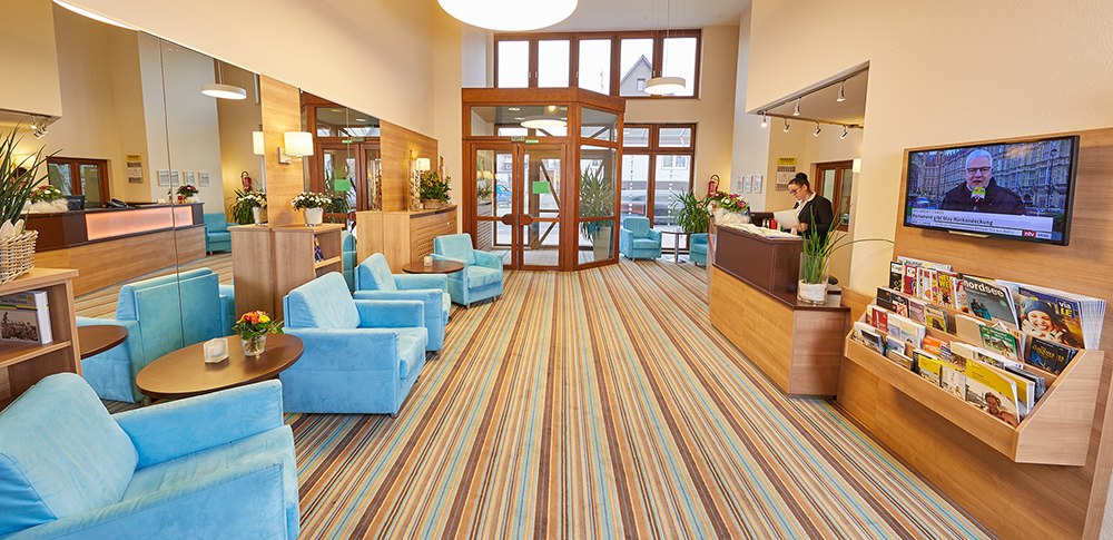 Helle Lobby mit blauen Sesseln, Holzmöbeln und gestreiftem Teppich. Rezeption mit Mitarbeiter, TV an der Wand, Zeitschriftenständer., © TOMAS