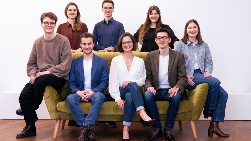 Acht junge Erwachsene posieren lächelnd um ein grünes Sofa vor einem weißen Hintergrund., © calens vocalensemble stuttgart Acht junge Erwachsene posieren lächelnd um ein grünes Sofa vor einem weißen Hintergrund., © calens vocalensemble stuttgart