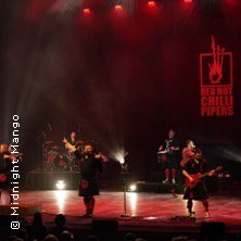 Die Red Hot Chilli Pipers performen auf einer Bühne, umgeben von rotem Licht. Dudelsäcke und Gitarren sind zu sehen., © links im Bild Die Red Hot Chilli Pipers performen auf einer Bühne, umgeben von rotem Licht. Dudelsäcke und Gitarren sind zu sehen., © links im Bild