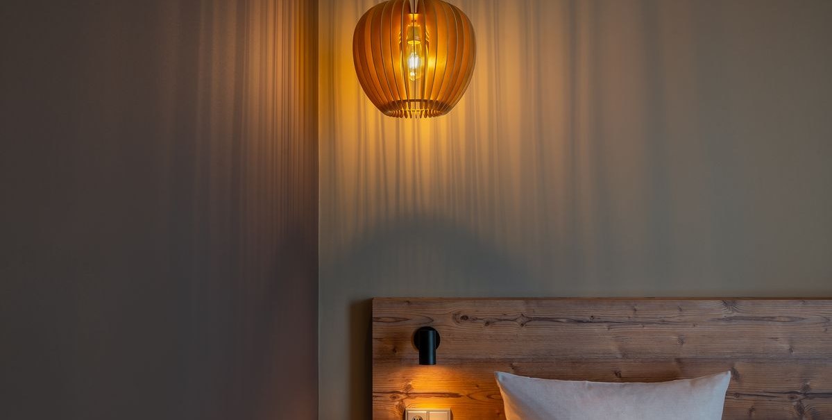 Eine hölzerne Lampe wirft warmes Licht auf eine Wand und ein Holzbett mit Kissen in einem modernen Zimmer., © TOMAS