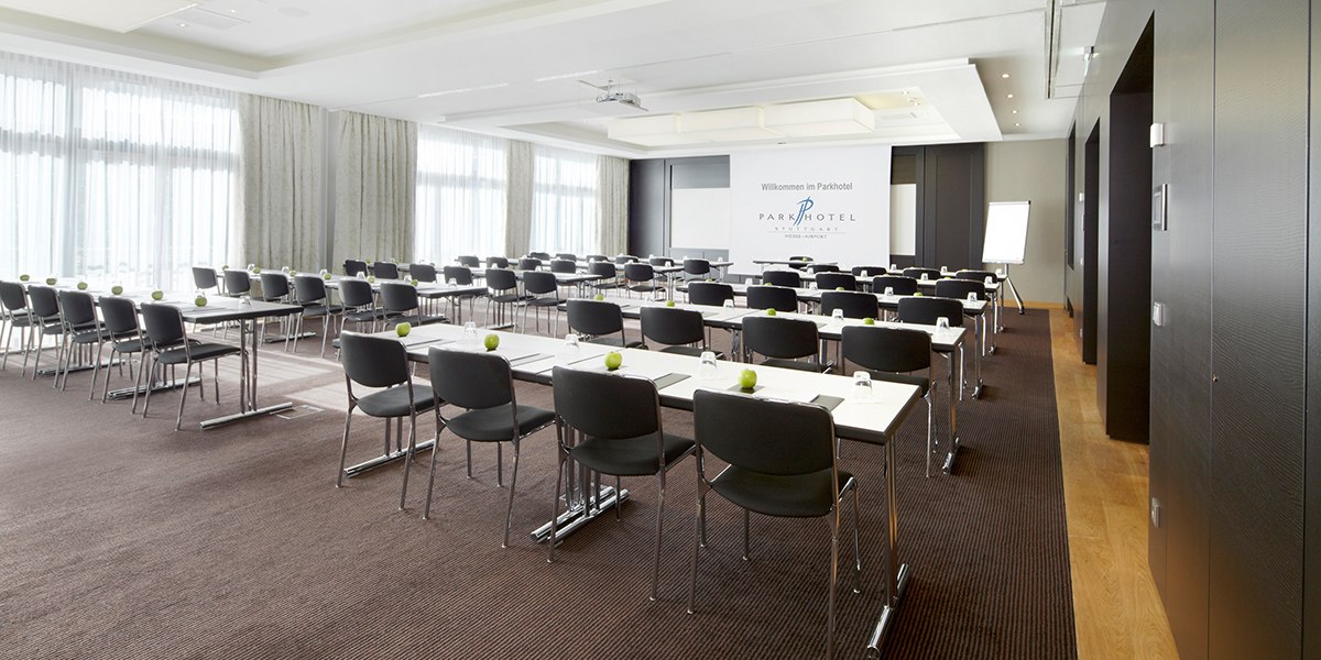 Ein moderner Konferenzraum mit Reihen von Tischen und Stühlen, einer Leinwand und einem Flipchart. Helle Vorhänge lassen Tageslicht herein., © Parkhotel Stuttgart Messe-Airport Ein moderner Konferenzraum mit Reihen von Tischen und Stühlen, einer Leinwand und einem Flipchart. Helle Vorhänge lassen Tageslicht herein., © Parkhotel Stuttgart Messe-Airport