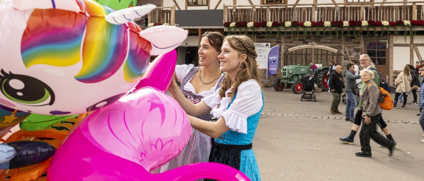 Two women in dirndls look at colorful balloons at a folk festival in Stuttgart. Half-timbered houses and a tractor can be seen in the background., © Stuttgart-Marketing GmbH, Sarah Schmid Two women in dirndls look at colorful balloons at a folk festival in Stuttgart. Half-timbered houses and a tractor can be seen in the background., © Stuttgart-Marketing GmbH, Sarah Schmid