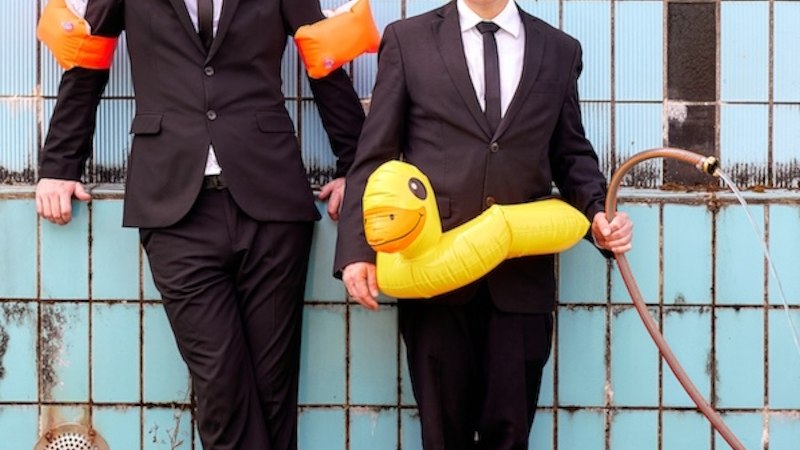 Zwei M&auml;nner in Anz&uuml;gen vor blauer Kachelwand. Einer tr&auml;gt Schwimmfl&uuml;gel, der andere h&auml;lt einen gelben Schwimmreifen und einen Wasserschlauch., &copy; Renitenztheater Stuttgart e.V.