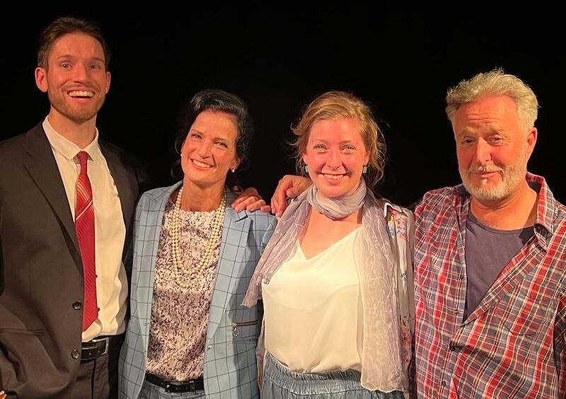 Four people pose smiling next to each other, two men and two women, in front of a dark background. They appear cheerful and relaxed., © Theater Atelier Four people pose smiling next to each other, two men and two women, in front of a dark background. They appear cheerful and relaxed., © Theater Atelier