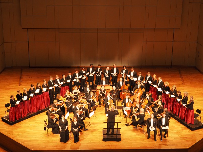 Ein Chor und Orchester auf einer Bühne. Der Dirigent steht vor den Musikern, die in formeller Kleidung auftreten. Der Konzertsaal ist gut beleuchtet., © Musik Podium Stuttgart