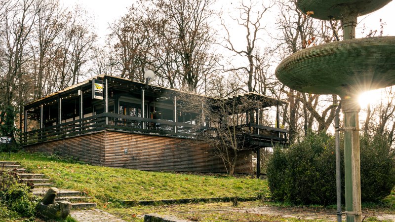 Geb&auml;ude mit Holzverkleidung und Terrasse in bewaldeter Umgebung, daneben ein gro&szlig;er, mehrstufiger Brunnen. Sonnenstrahlen scheinen durch die B&auml;ume., &copy; Stuttgart Marketing GmbH, Sarah Schmid