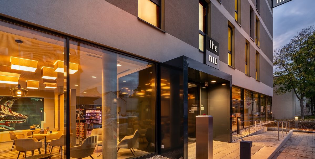 Moderner Hoteleingang mit Glasfassade, beleuchtetem Innenbereich und dem Schriftzug 'the niu'. Au&szlig;enbereich mit Gehweg und Pflanzen., &copy; Novum Hospitality