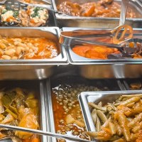 Ein Buffet mit verschiedenen warmen Speisen in Edelstahlbehältern, darunter Bohnen, Gemüse und gefüllte Auberginen., © Zauberküche, Stuttgart