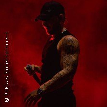 Ein tätowierter Mann in schwarzem Tanktop und Kappe steht mit Mikrofon vor einem dramatisch roten Hintergrund., © links im Bild Ein tätowierter Mann in schwarzem Tanktop und Kappe steht mit Mikrofon vor einem dramatisch roten Hintergrund., © links im Bild