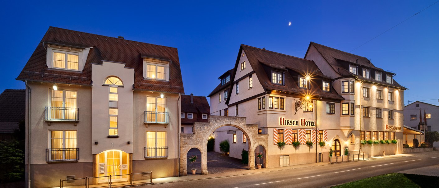 Ein Hotel bei Nacht mit beleuchteten Fenstern und einem Torbogen. Der Himmel ist dunkelblau, der Mond ist sichtbar. Das Gebäude wirkt einladend und gepflegt., © Hirsch Hotel