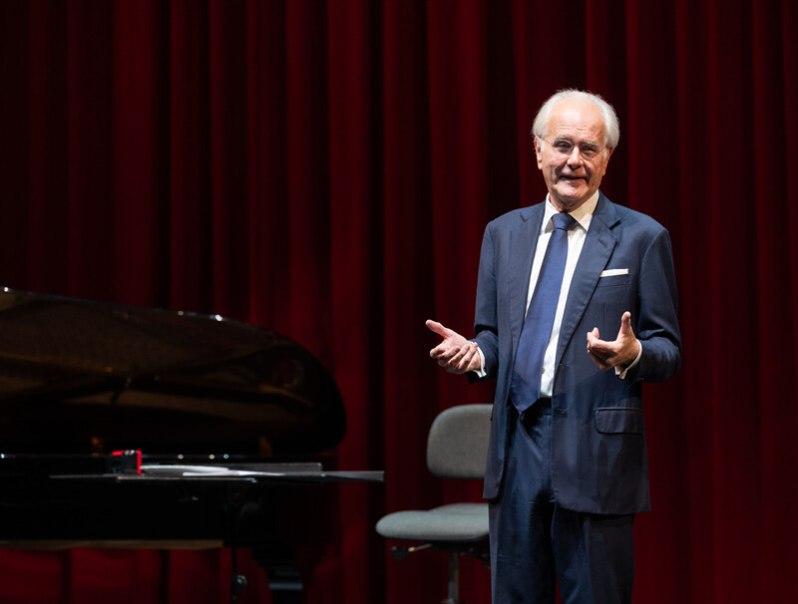 Ein Mann im Anzug spricht auf einer Bühne vor einem roten Vorhang, neben einem Flügel., © Württembergische Staatstheater Stuttgart