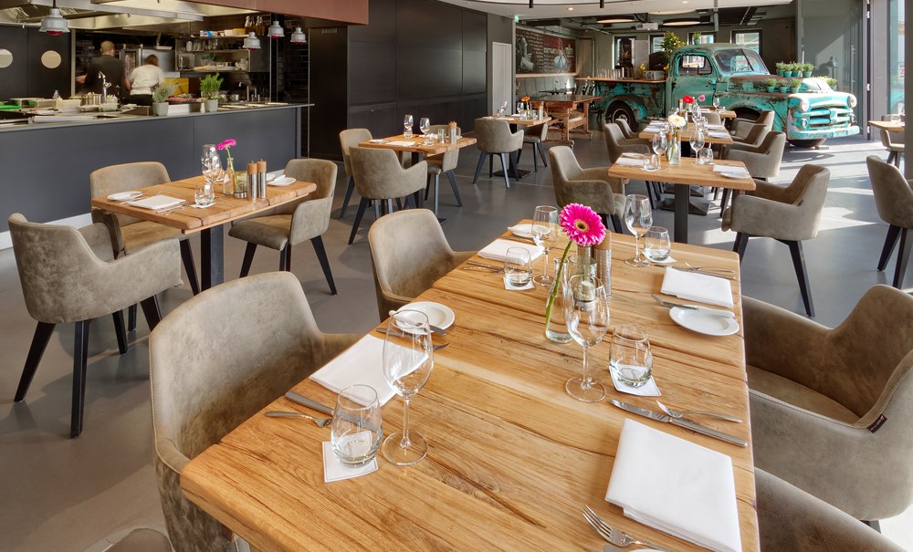 Ein modernes Restaurant mit Holztischen, stilvoll gedeckt. Im Hintergrund steht ein alter grüner Truck als Dekoration., © V8 HOTEL Motorworld Region Stuttgart