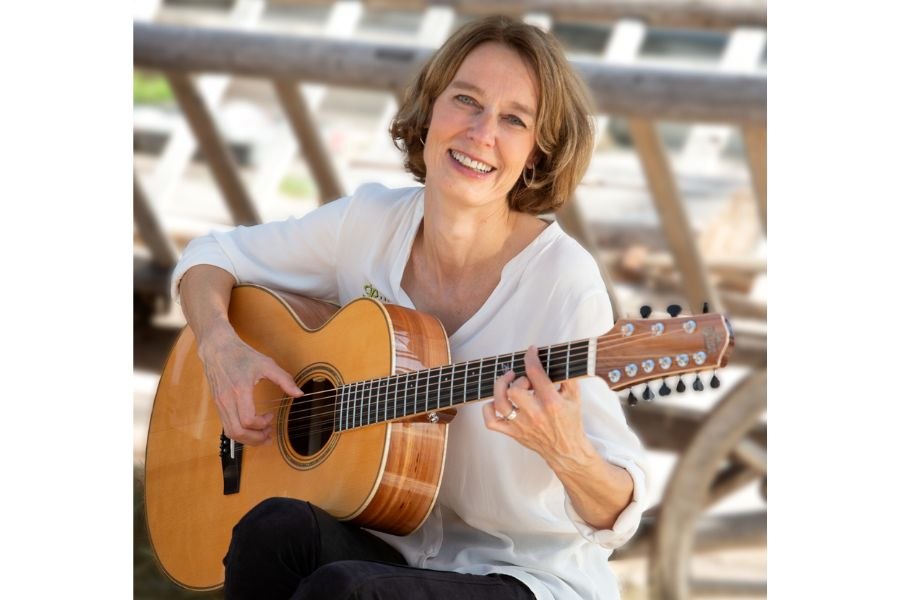 Eine Frau sitzt draußen und spielt Gitarre, während sie in die Kamera lächelt. Sie trägt ein weißes Hemd und wirkt entspannt und glücklich., © Sandra Wolf Eine Frau sitzt draußen und spielt Gitarre, während sie in die Kamera lächelt. Sie trägt ein weißes Hemd und wirkt entspannt und glücklich., © Sandra Wolf