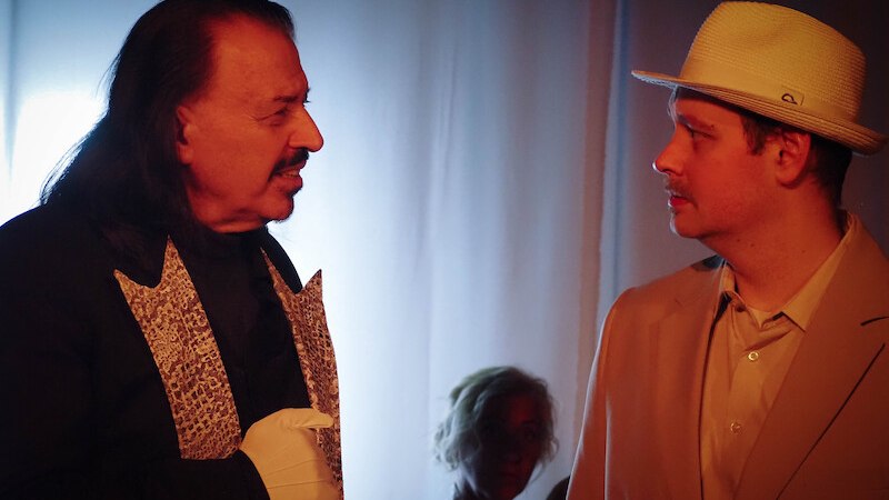 Zwei M&auml;nner in einem Theaterst&uuml;ck, einer tr&auml;gt einen eleganten Anzug mit Handschuhen, der andere einen hellen Anzug mit Hut. Im Hintergrund eine Frau., &copy; Theater Atelier