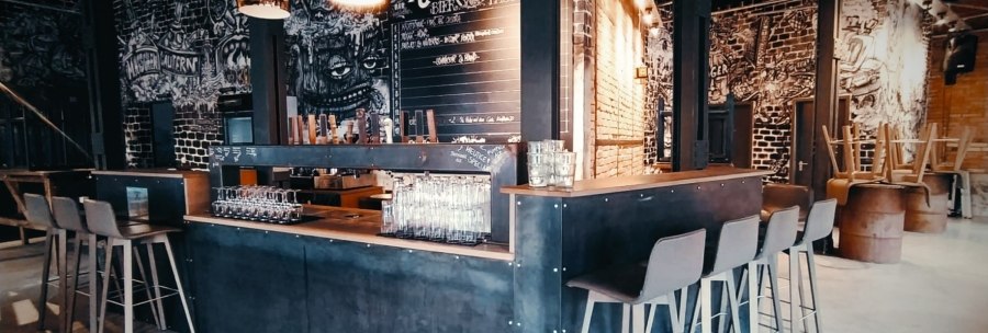 Modern bar with industrial design, high stools and wall art. Glasses are placed on the counter, in the background a table with drinks., © Kraftpaule Modern bar with industrial design, high stools and wall art. Glasses are placed on the counter, in the background a table with drinks., © Kraftpaule
