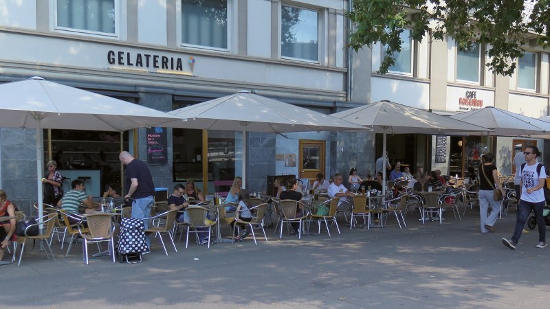 People sit under parasols in front of the Kaiserbau gelateria on Marienplatz. A few passers-by walk past., © Stuttgart-Marketing GmbH