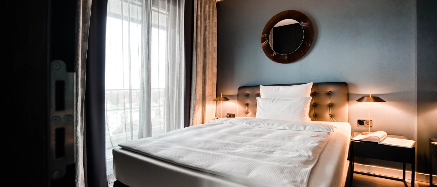 Modernes Hotelzimmer mit großem Bett, runden Spiegel an der Wand, Nachttischlampen und bodenlangen Vorhängen vor einem Fenster., © CLOUD N°7 APARTMENTS GmbH Modernes Hotelzimmer mit großem Bett, runden Spiegel an der Wand, Nachttischlampen und bodenlangen Vorhängen vor einem Fenster., © CLOUD N°7 APARTMENTS GmbH