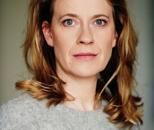A woman with blonde hair and a gray sweater looks directly into the camera. The background is neutral., &copy; Theaterhaus Stuttgart e.V.