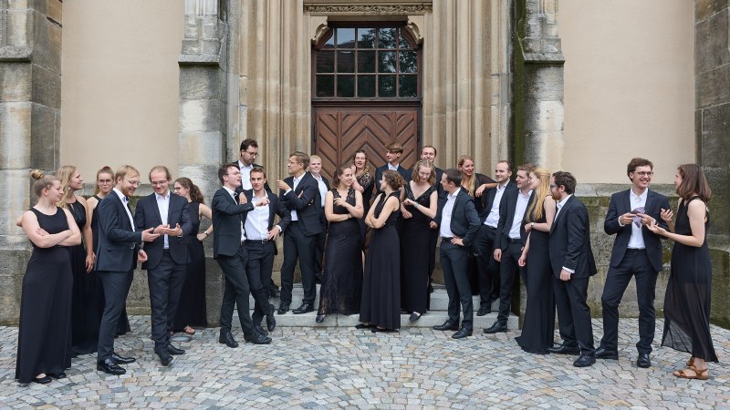 Eine Gruppe von Menschen in eleganter Abendkleidung steht vor einem alten Gebäude mit verzierten Türen und Steinmauern., © collegium vocale Stuttgart Eine Gruppe von Menschen in eleganter Abendkleidung steht vor einem alten Gebäude mit verzierten Türen und Steinmauern., © collegium vocale Stuttgart