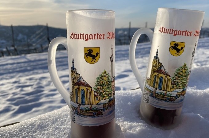 Zwei Glühweintassen mit Stuttgart-Motiv stehen im Schnee. Im Hintergrund sind Weinberge und ein blauer Himmel zu sehen., © Weingenuss Stuttgart