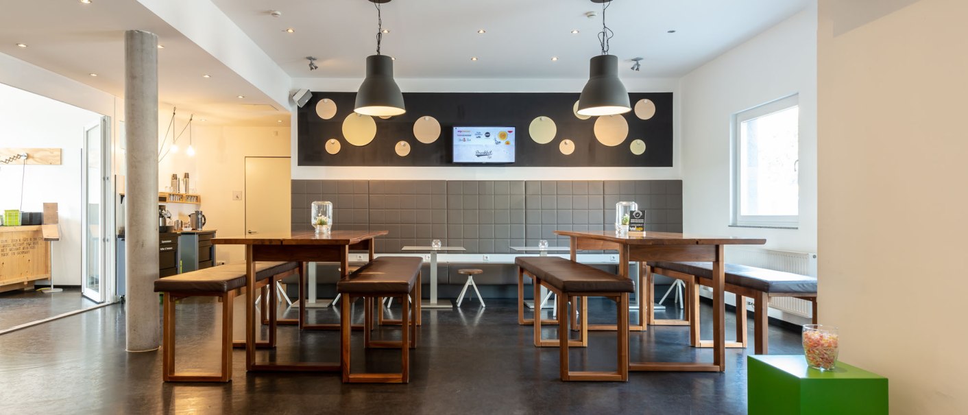 Modern lobby with wooden tables and benches, decorative lamps and a wall with circular motifs. A green stool with a jar of sweets stands in the foreground., &copy; a2 HOTELS Plochingen
