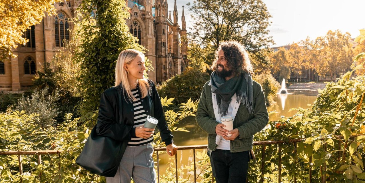 Two people stand smiling in front of a church and a pond, surrounded by autumnal foliage. They are holding coffee cups in their hands., &copy; SMG, Sarah Schmid