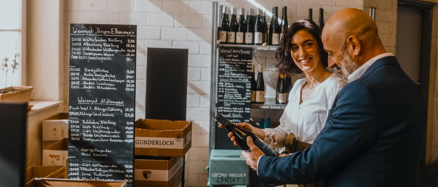 Zwei Personen in einer Weinhandlung, eine Frau lächelt, während sie eine Weinflasche hält. Im Hintergrund sind Weine und eine Preistafel zu sehen., © SMG, Alwin Maigler